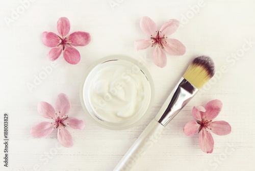 Beauty skincare cream in cosmetic jar, delicate fresh pink spring blossom, application brush, top view white wooden table.