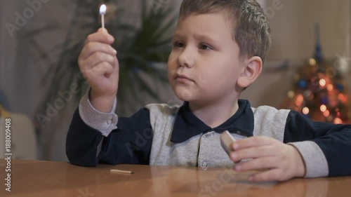 Little boy holding a safety match and looking at the flame. New year tree background. Dangerous toy in kids hands photo