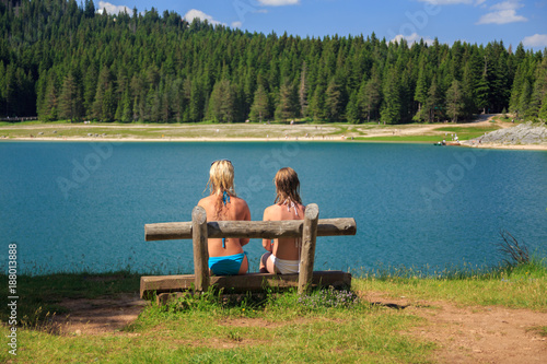 rear view of two girls sitting on a bench near the lake in the mountains