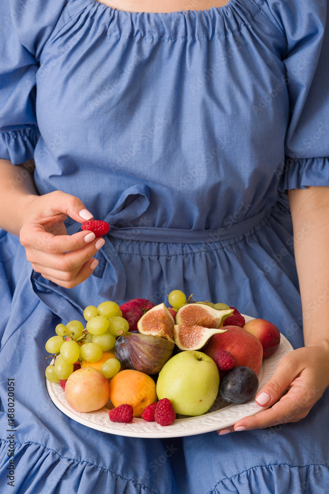 woman sitting cozy on the couch and enjoying delicious summer fruit and berries. Girl holding white plate with apricots, raspberries, peaches and figs on her knees. Healthy eating.