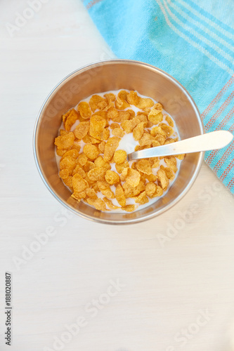 Cornflakes in a metal bowl with milk on a painted white wooden background. Composition with a dish towel and spoon.
