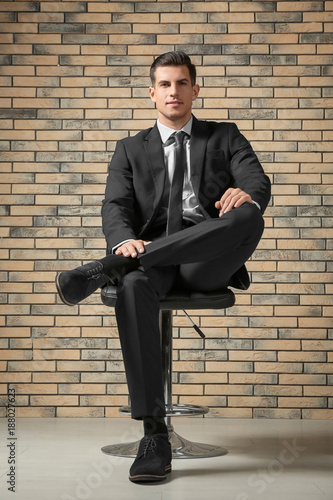 Handsome man in elegant suit sitting on chair near brick wall © Africa Studio