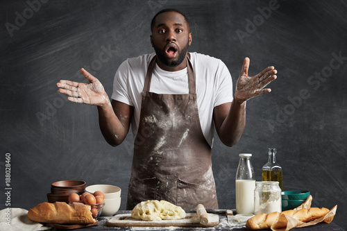 Uncertain black man shrugs shouldres as stands at kitchen table, being hesitant what ingridient to put in his dish, isolated over black chalk background. People, ethnicity and culinary concept photo