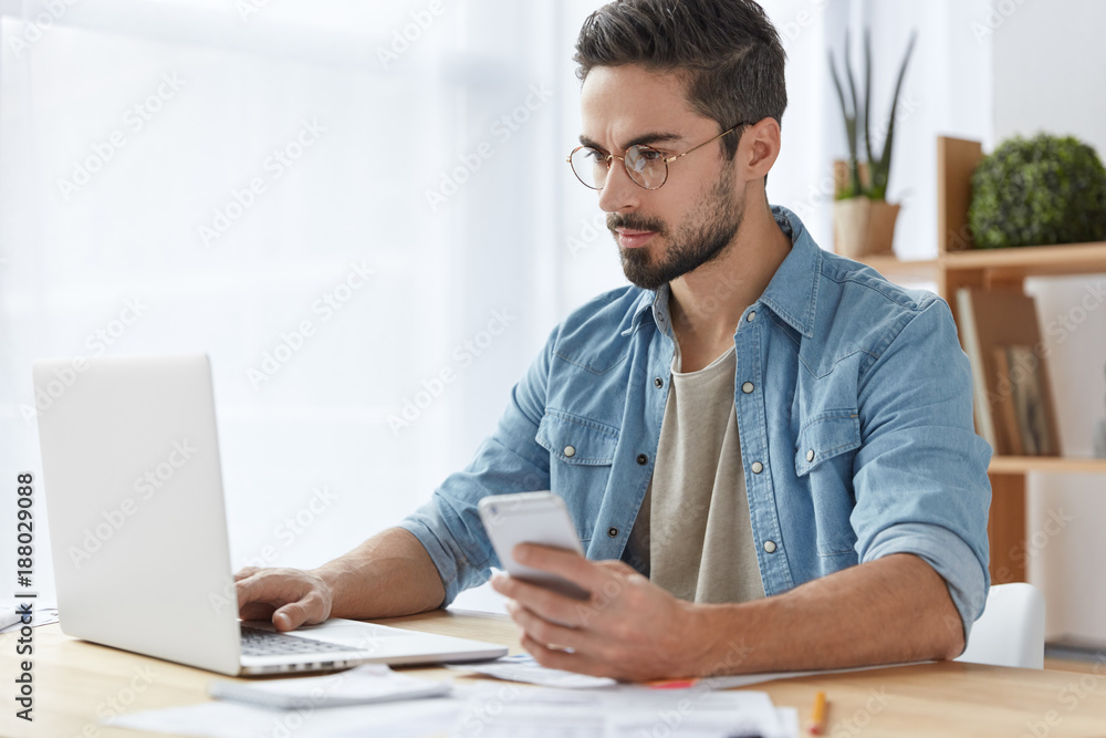 Photo of serious focused man with beard and mustache uses modern digital smart phone device, chats in social networks, keyboards on laptop computer, reads news on websites, makes payment online