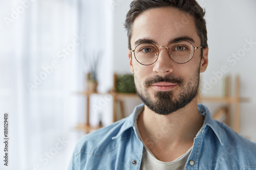 Close up portrait of handsome bearded man wears round spectacles, has appealing appearance with beard and mustache, dressed in fashionable clothes, stands against cozy interior. Student poses indoor