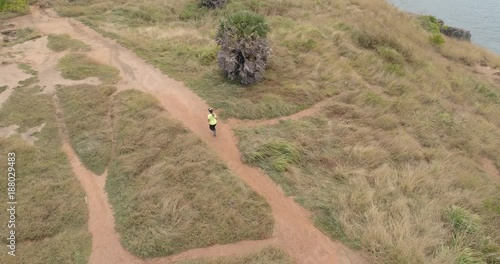 Video from drone. Sportive beautiful girl running by rocky road at cliff washed by sea on a sunny day. The woman run between rocks. Hiking. Outdoor taining. Nai Harn cliff, Phuket island, Thailan. photo