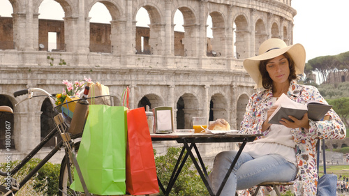 Canvas Print Young woman tourist reads a book sitting at the table outside a bar restaurant in front of the Colosseum in Rome with coffee, juice and cornetto