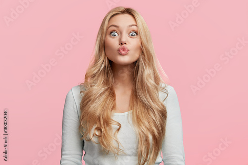 Φωτογραφία Portrait of adorable blonde woman pouts lips and foolishes alone, makes grimace, rounds lips for making kiss, isolated over pink background