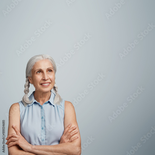 Pleasant looking cheerful grandmother keeps arms folded, demonstrates shining smile, looks thughtfully upwards, remembers pleasant moments spent with children and grandchildren, blank copy space