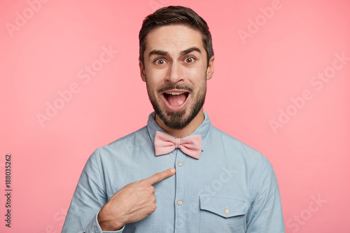 Puzzled young male model indicates at himself with indignant expression, demonstrates new bowtie, isolated over pink background. Handsome man looks in bewilderment at camera, gestures indoor