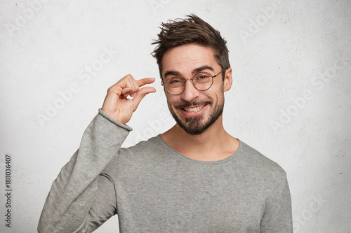 Positive handsome man with stylish hairdo, dressed in casual sweater, shows something very tiny or small, being in good mood, poses against white concrete background. Young fashionable male gestures photo