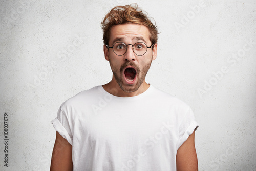 Photo of amazed fashionable young male model expresses great wonder and surprisment after hearing shocking news, dressed in white casual t shirt, isolated over concrete wall. Amazement concept