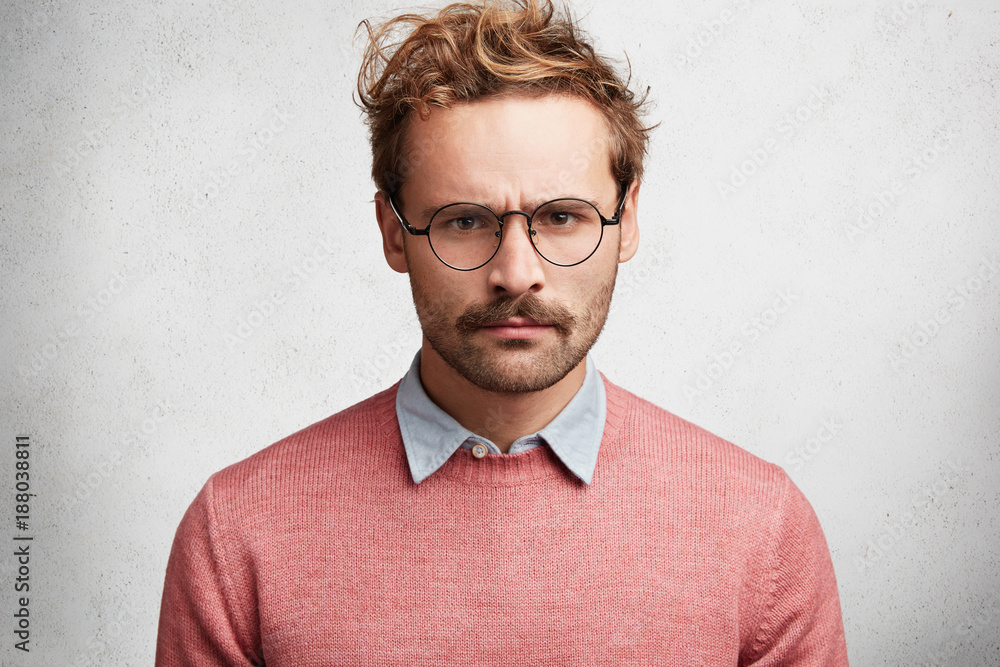 Fototapeta premium Horizontal shot of grumpy angry man frowns face in dissatisfaction, looks through spectacles, has gloomy expression, poses against white concrete background. Discontent displeased young human male
