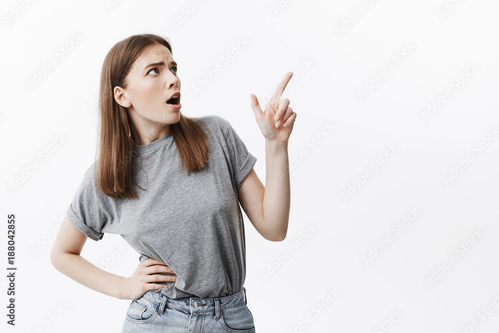Negative emotions. Portrait of expressive young good-looking student girl iwith dark hair in grey clothes looking aside with opened mouth and mean expression, pointing on white wall with finger. Copy Negative emotions. Portrait of expressive young good-looking student girl iwith dark hair in grey clothes looking aside with opened mouth and mean expression, pointing on white wall with finger. Copy
