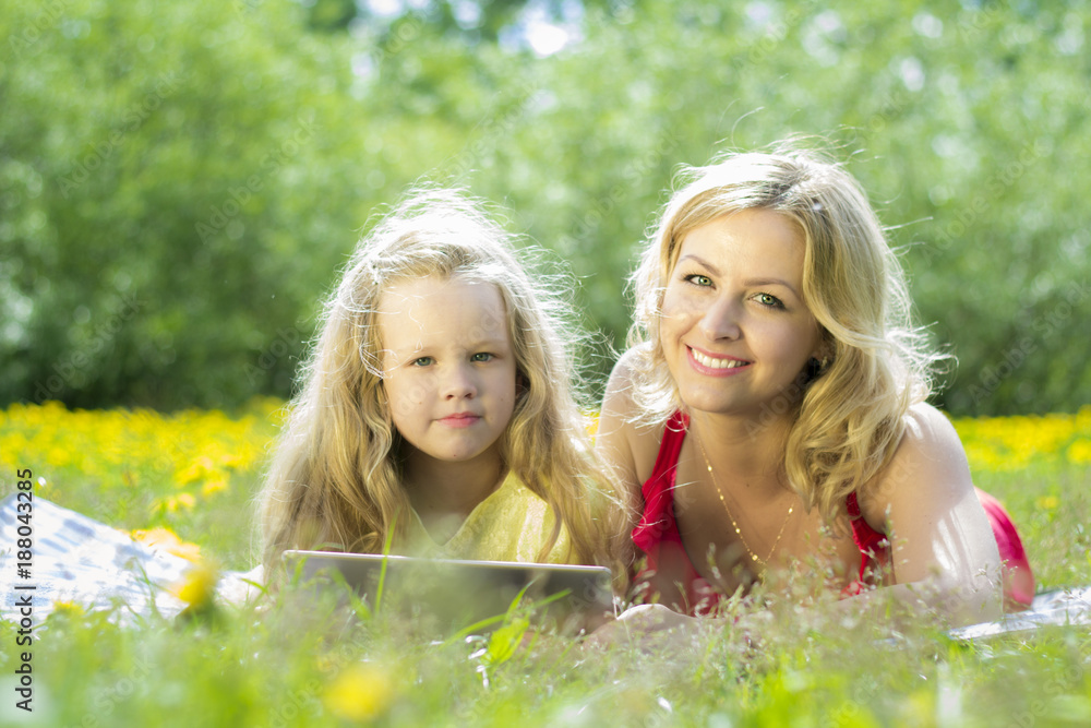 Beautiful woman and girl playing on a tablet, in the nature, lying on a blanket in the grass. Beautiful woman and girl playing on a tablet, in the nature, lying on a blanket in the grass.