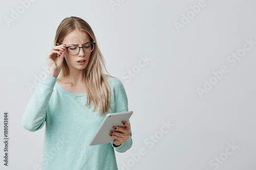 Attractive blonde female manager in stylsh eyewear works, look nervously at screen of tablet finds out unpleasant news. Annoyed Caucasian woman dressed in blue sweater uses modern technologies. photo