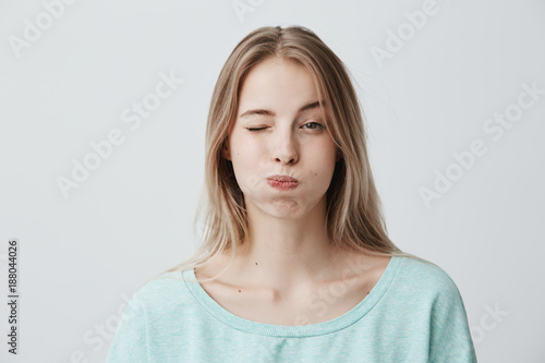 Portrait of frowning blonde young woman dressed in light blue sweater, pouting her cheeks, blinking, doesn`t want to share information. Human emotions, face expression, reaction and attitute