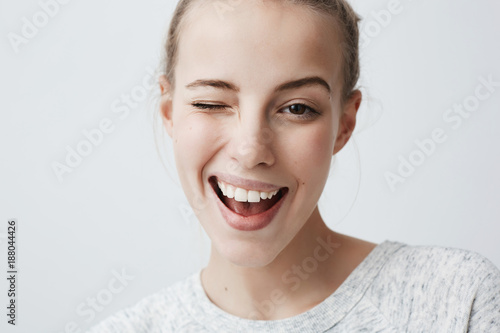 Obraz na plátne Close-up studio shot of positive coquettish young European woman with dark eyes, smiling happily, blinking at camera in a playful manner, flirting with you