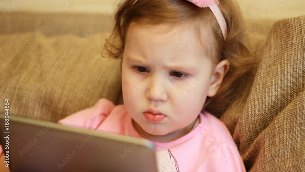 A little cute girl is sitting in sofa in a living room, looking cartoon and playing the game on a tablet.. Funny child looks on the computer screen and plays downloaded application. Close-up portrait A little cute girl is sitting in sofa in a living room, looking cartoon and playing the game on a tablet.. Funny child looks on the computer screen and plays downloaded application. Close-up portrait