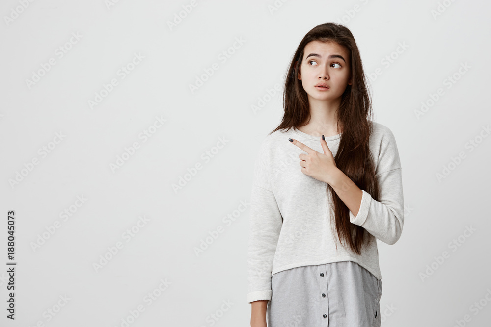 Cute cheerful brunette young female pointing with index finger away, showing something interesting and exciting on studio wall with copy space for your text or advertising content Cute cheerful brunette young female pointing with index finger away, showing something interesting and exciting on studio wall with copy space for your text or advertising content