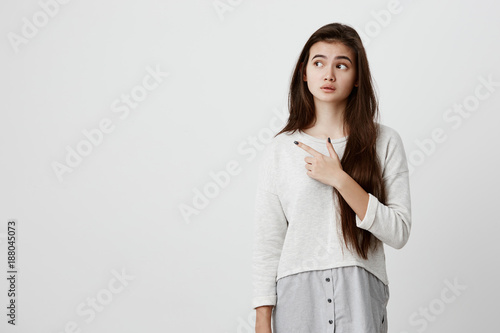 Cute cheerful brunette young female pointing with index finger away, showing something interesting and exciting on studio wall with copy space for your text or advertising content