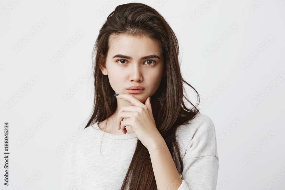 Human emotions, feelings, reaction and attitude. Beautiful girl in casual t-shirt with long dark straight hair, keeping hand on chin in doubt and suspicion, feeling sceptical about something Human emotions, feelings, reaction and attitude. Beautiful girl in casual t-shirt with long dark straight hair, keeping hand on chin in doubt and suspicion, feeling sceptical about something