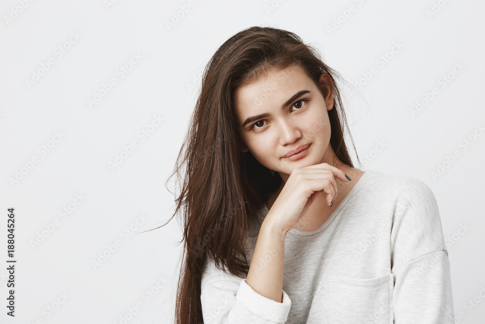 Young brunette female with oval face, dark appealing eyes having hair loose dressed in long-sleeved t-shirt holding hand under chin looking with dreamy expression at camera, thinking about something Young brunette female with oval face, dark appealing eyes having hair loose dressed in long-sleeved t-shirt holding hand under chin looking with dreamy expression at camera, thinking about something