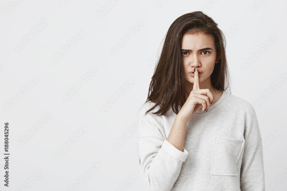 Portrait of emotional brunette girl dressed casually holding index finger on lips, saying 'shh', asking to keep silence or to keep her secret. Face expressions, emotions and feelings, body language Portrait of emotional brunette girl dressed casually holding index finger on lips, saying 'shh', asking to keep silence or to keep her secret. Face expressions, emotions and feelings, body language
