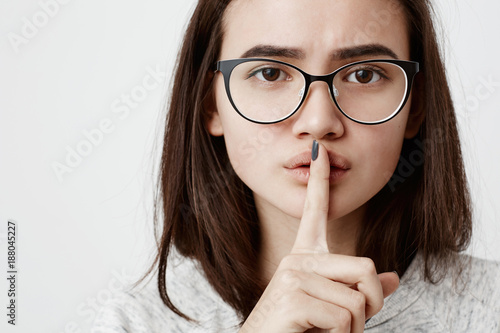 Worried serious dark-haired teenage girl wearing casual t-shirt and stylish spectacles holding index finger on her lips, asking to keep silence and not to make noize. Human face expressions