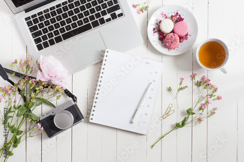Feminine flat lay workspace with laptop  cup of tea  planner  macarons. retro camera and flowers on white wooden table. Top view mock up.