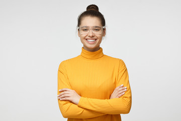 Portrait of smiling attractive female in mustard yellow sweater and transparent eyeglasses, standing with crossed arms isolated on gray background