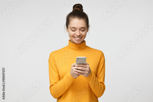 Wallpaper Mural Yong pretty female standing isolated on gray background looking attentively at screen of cellphone, browsing web pages and smiling nicely while chatting Torontodigital.ca