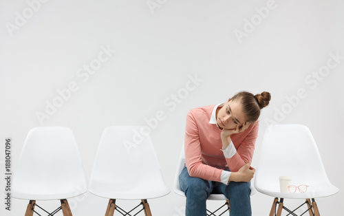 Tired and bored, exhausted female in casual pink sweater sitting in the line and holding head with one hand, waiting for something