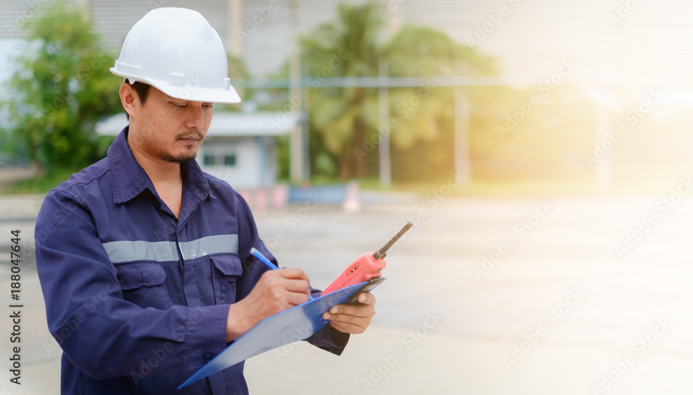 Fototapeta premium Asian engineer in safety uniform and white helmet taking note on clipboard on blurred industry plant background