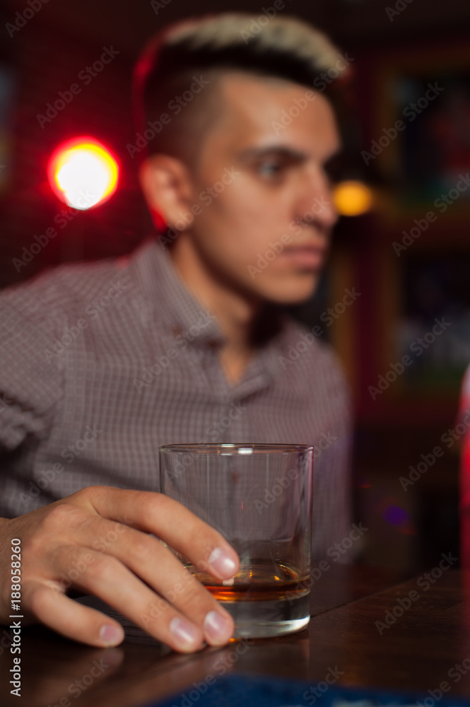 Blurry young hipster man sits at bar counter and holds glass of whiskey after long working day Blurry young hipster man sits at bar counter and holds glass of whiskey after long working day