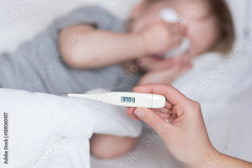 Close-up thermometer with fever heat 38. Mother measuring temperature of her ill kid at background. Sick child with high fever lying in bed at home. Mom Hand with thermometer close up