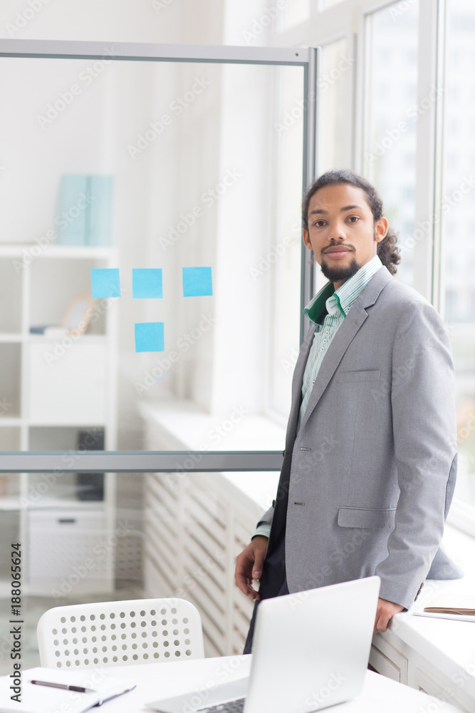 Serious businessman in elegant suit looking at camera while standing by window in office