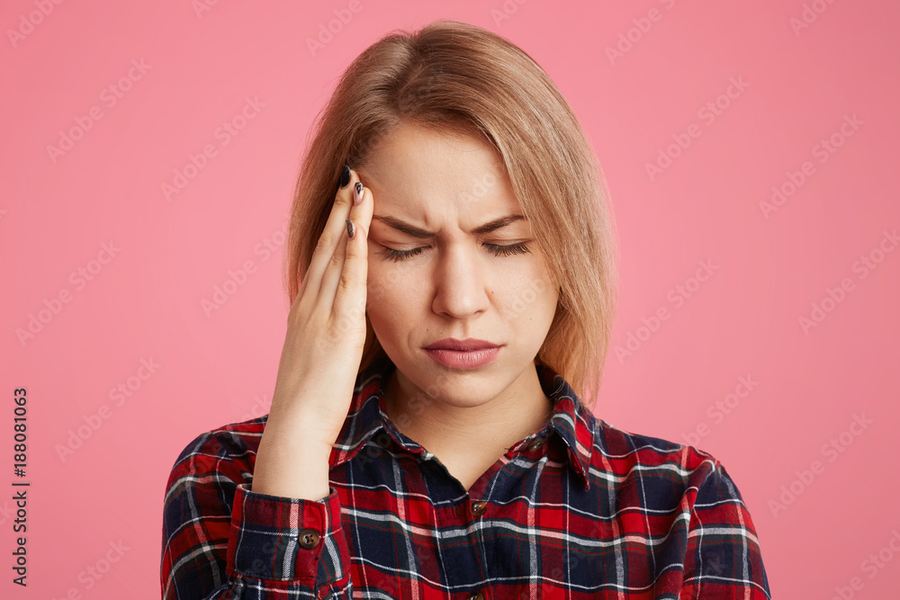 Close up portrait of stressful beautiful female student has headache after long exam preparation, closes eyes and scratches head, tries to overcome terrible pain, isolated over pink background Close up portrait of stressful beautiful female student has headache after long exam preparation, closes eyes and scratches head, tries to overcome terrible pain, isolated over pink background