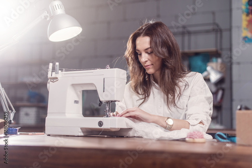 Attractive Caucasian seamstress working stitching with sewing machine at her workplace in studio loft interior