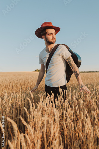 Young musician in hat with guitar walking on a summer wheat field. Bearded stylish young man. © stone36