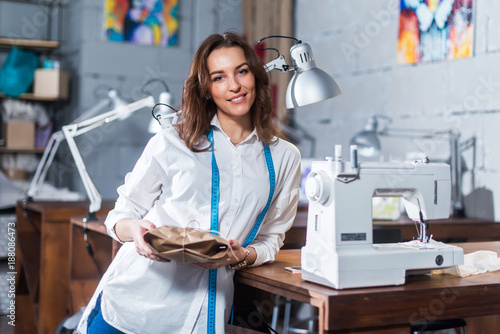 Portrait of smiling European fashion designer standing next to sewing machine holding a gift packed in craft paper in studio