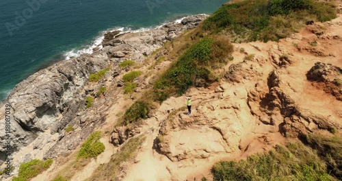 Video from drone. Sportive beautiful girl running by rocky road at cliff washed by sea on a sunny day. The woman run between rocks. Hiking. Outdoor taining. Nai Harn cliff, Phuket island, Thailand photo