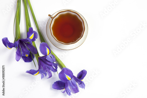 Fototapeta Naklejka Na Ścianę i Meble -  Violet Irises xiphium (Bulbous iris, sibirica) with cup of tea on white background with space for text. Top view, flat . Holiday greeting card for Valentine's Day, Woman's Day, Mother's Day, Easter!