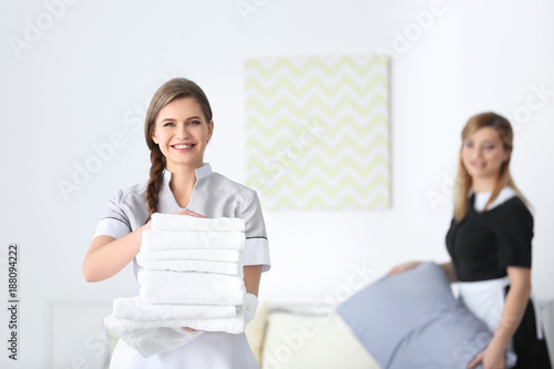 Young chambermaid in uniform holding stack of clean towels indoors