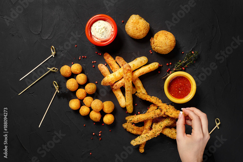 Mushrooms in batter, cheese balls, cheese sticks and cheese rings in batter, on black background, with herbs, sauce, ketchup and pepper. Top view