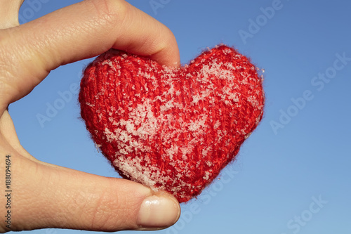 red knitted heart is covered with cold ice crystals lies in the warm fingers on blue sky background