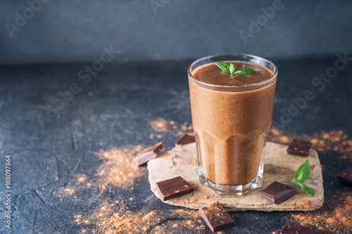 Chocolate smoothie with banana, decorated with mint leaf on the dark background with pieces of chocolate and cocoa powder. Healthy diet food. Selective focus, space for text.