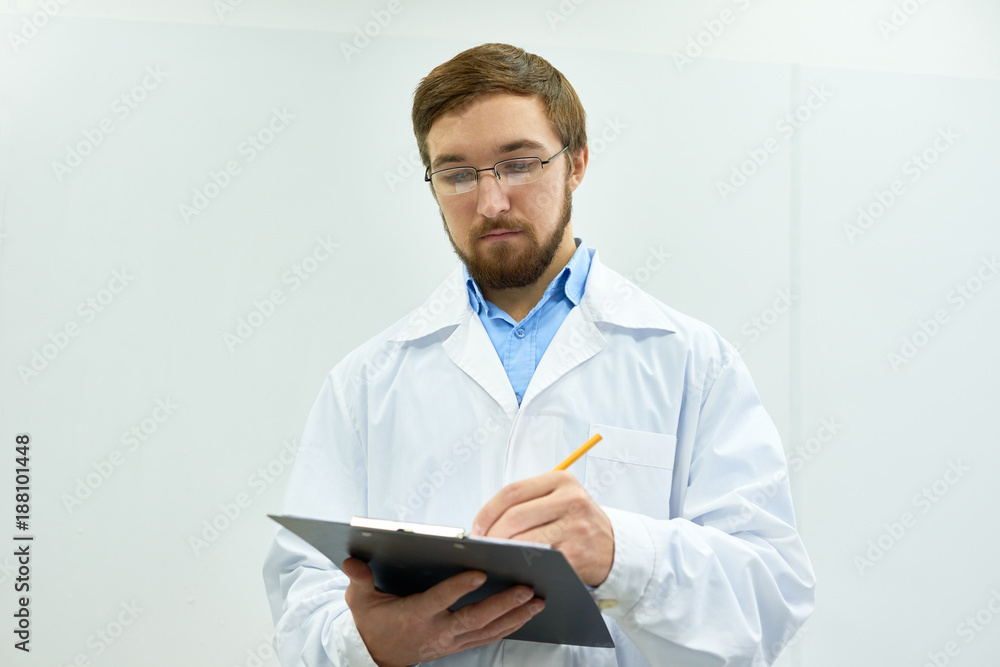 Portrait of young bearded doctor wearing glasses standing against white wall writing on clipboard, copy space Portrait of young bearded doctor wearing glasses standing against white wall writing on clipboard, copy space