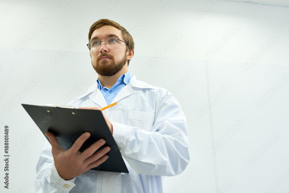 Low angle  portrait of young bearded doctor wearing glasses standing against white wall writing on clipboard, copy space