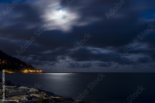Seascape at night with moon reflected in the sea. Deep blue night sky with beautiful clouds. Illuminated village on the coast.